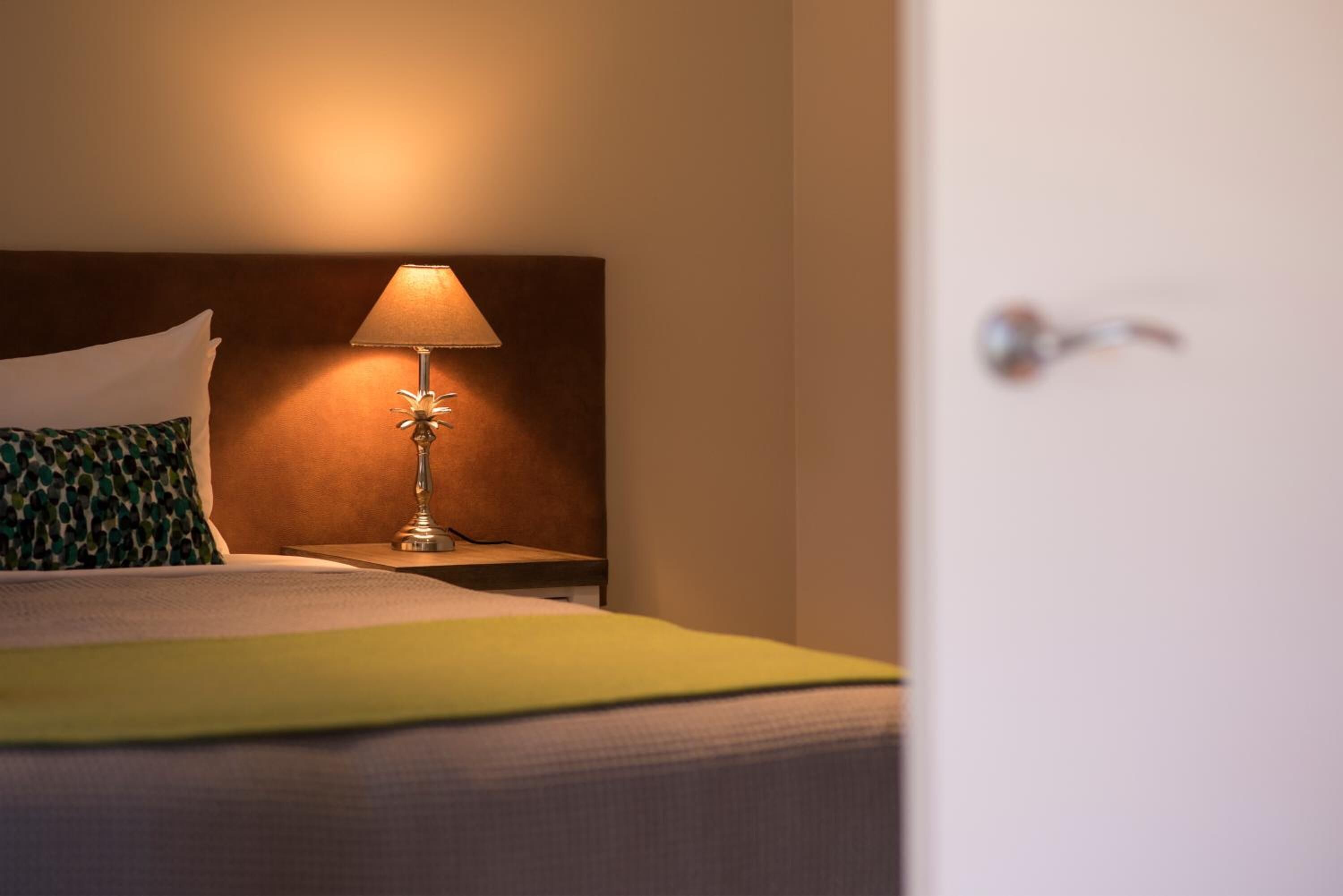 Ett hotellrum med en säng, ett nattduksbord med en lampa och en dörr.