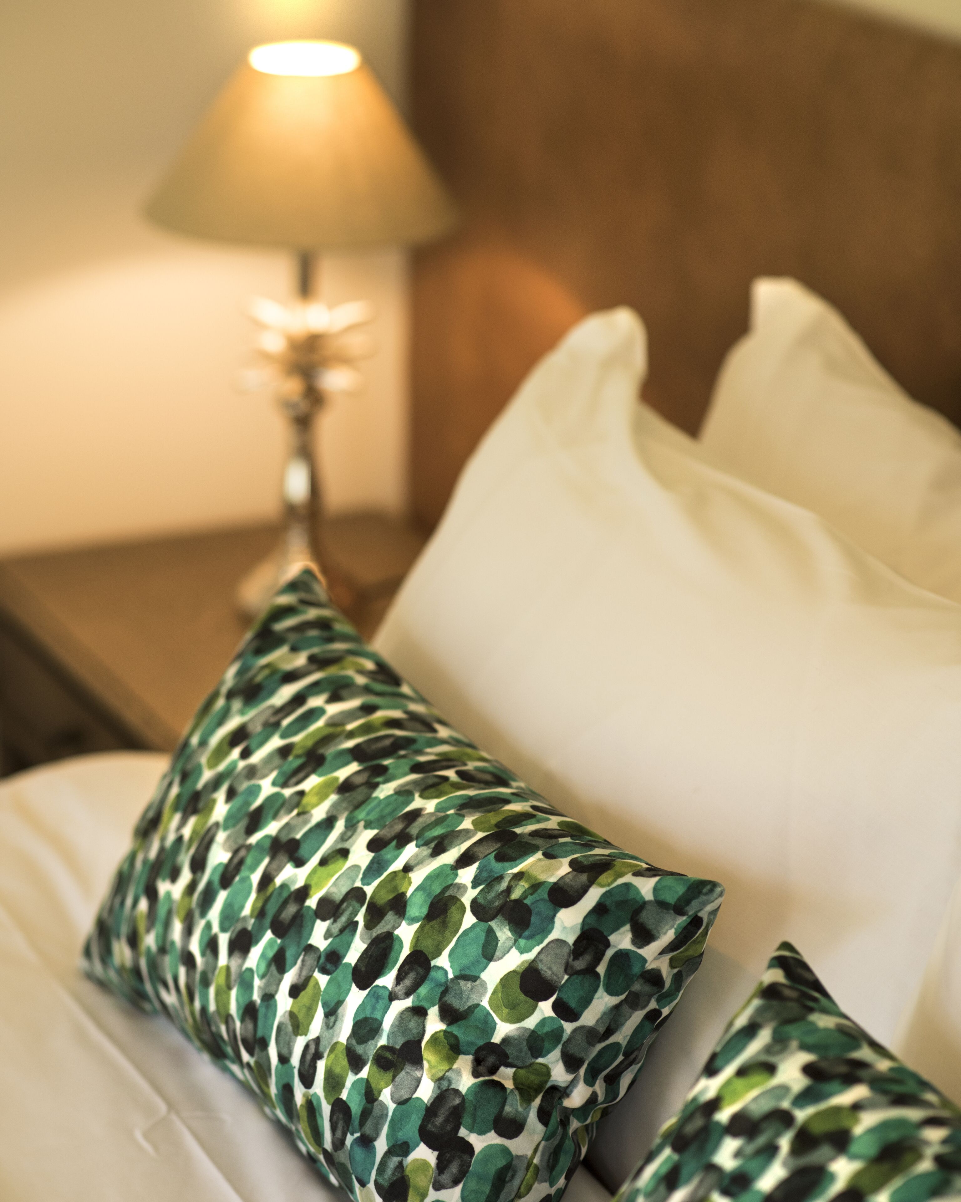Siististi pedattu sänky, jossa on valkoisia ja kuviollisia tyynyjä, yöpöytälamppu ja puinen yöpöytä.