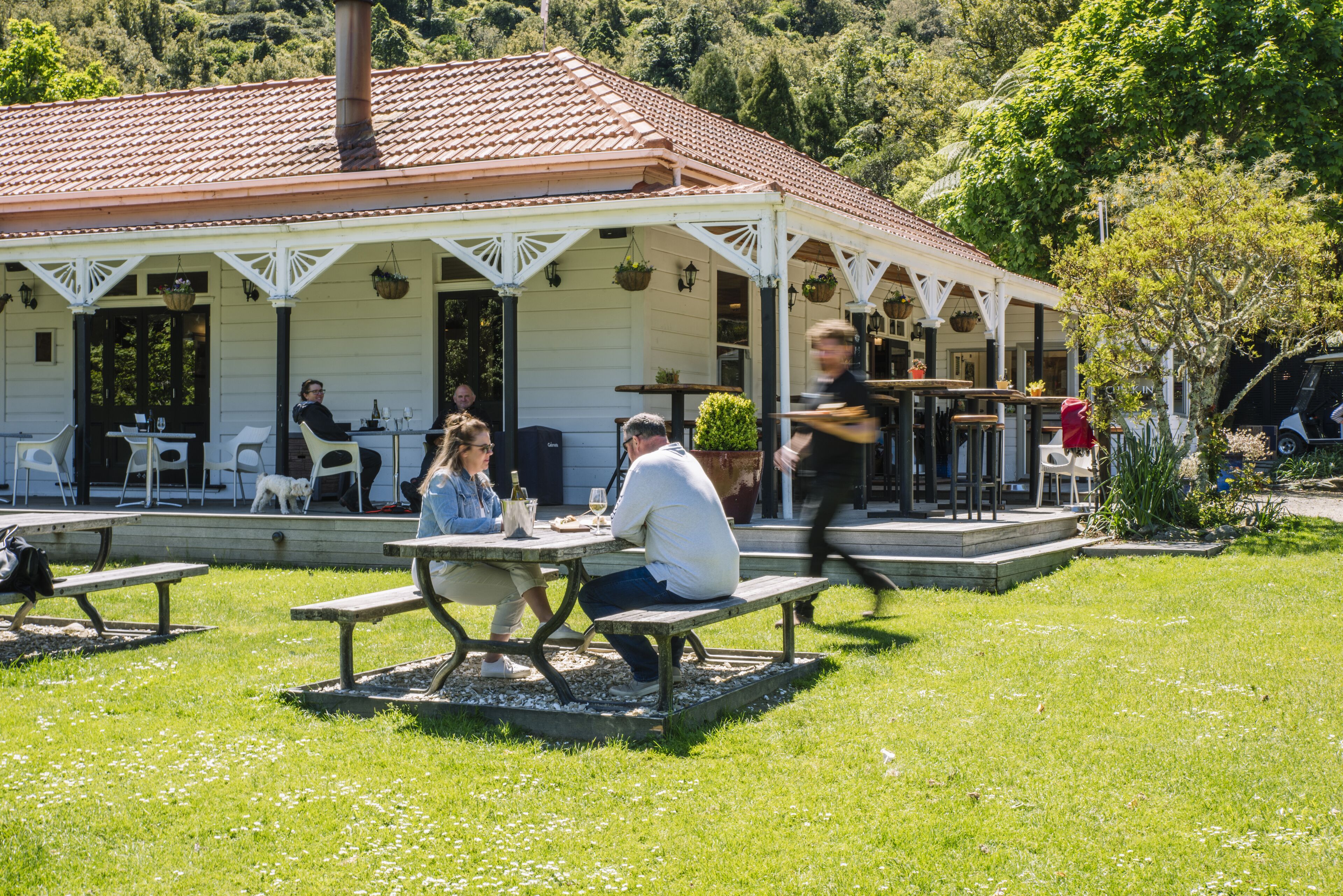 An outdoor cafe with tables and chairs, a covered seating area, and a building with a tiled roof.