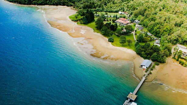 An aerial view of a coastal area with a pier extending into the sea, surrounded by lush greenery and a few buildings. - Furneaux Lodge (Endeavour Inlet)