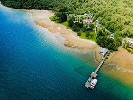 Vista aérea de uma área costeira com um cais que se estende para o mar, rodeada por vegetação exuberante e alguns edifícios.