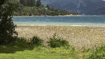 Una playa con costa rocosa, un primer plano de césped y montañas y árboles al fondo.