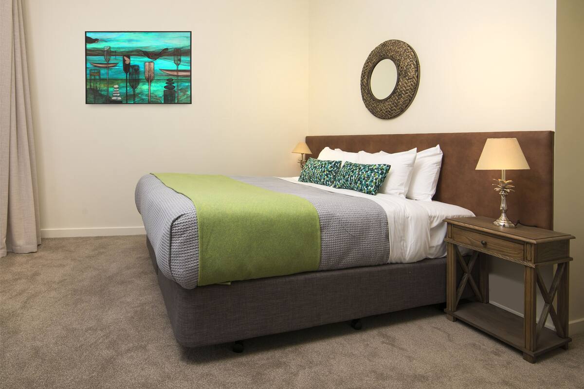 A neatly made bed with a green and gray blanket, a wooden nightstand with a lamp, and a wall-mounted mirror.