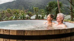 A hot tub with a wooden frame, surrounded by lush greenery and a mountainous backdrop. - Furneaux Lodge (Endeavour Inlet)