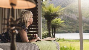A person reading a book in a room with a view of a lake and mountains. - Furneaux Lodge (Endeavour Inlet)