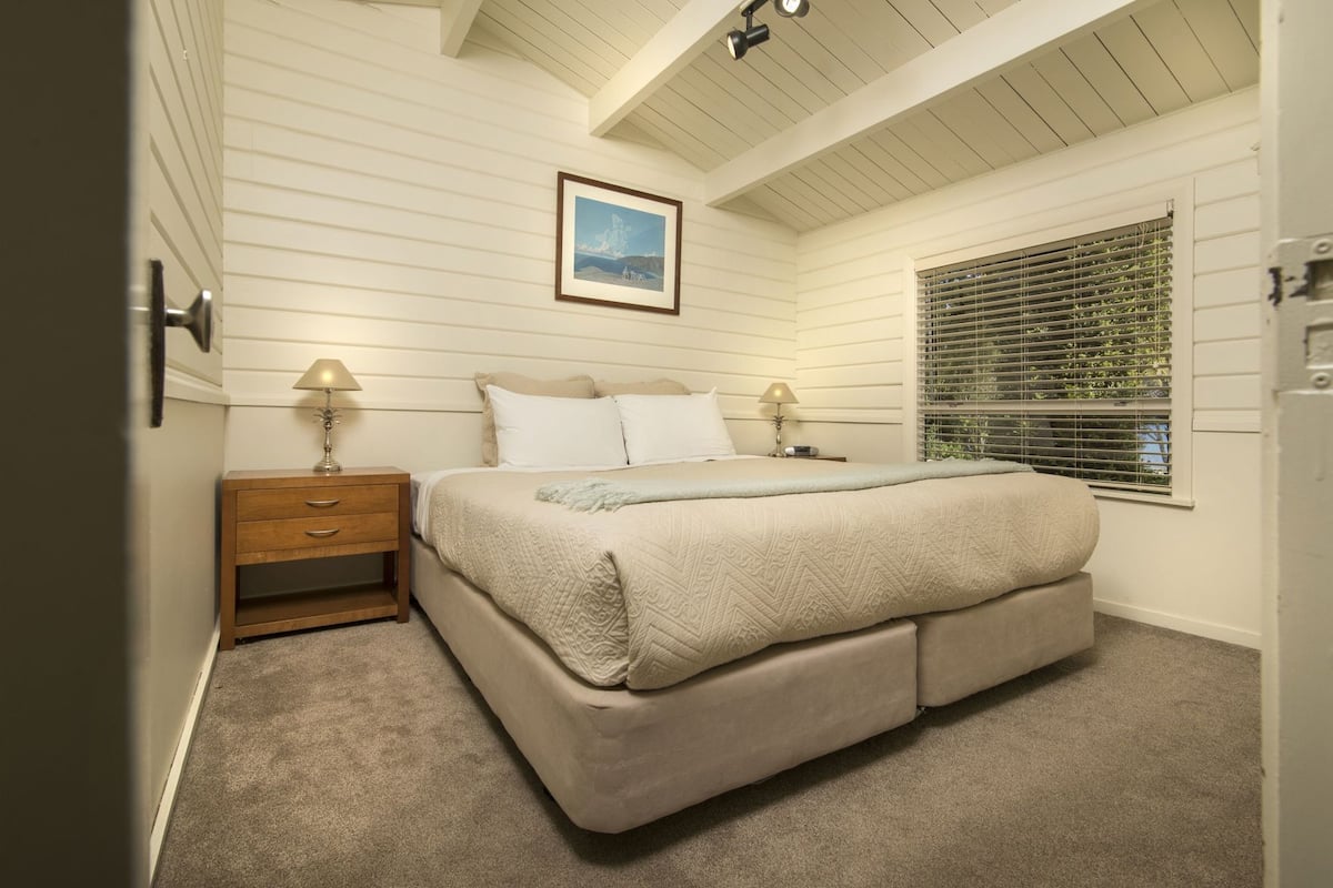 A bedroom with a bed, two bedside tables, a window with blinds, a ceiling light, and a framed picture on the wall.