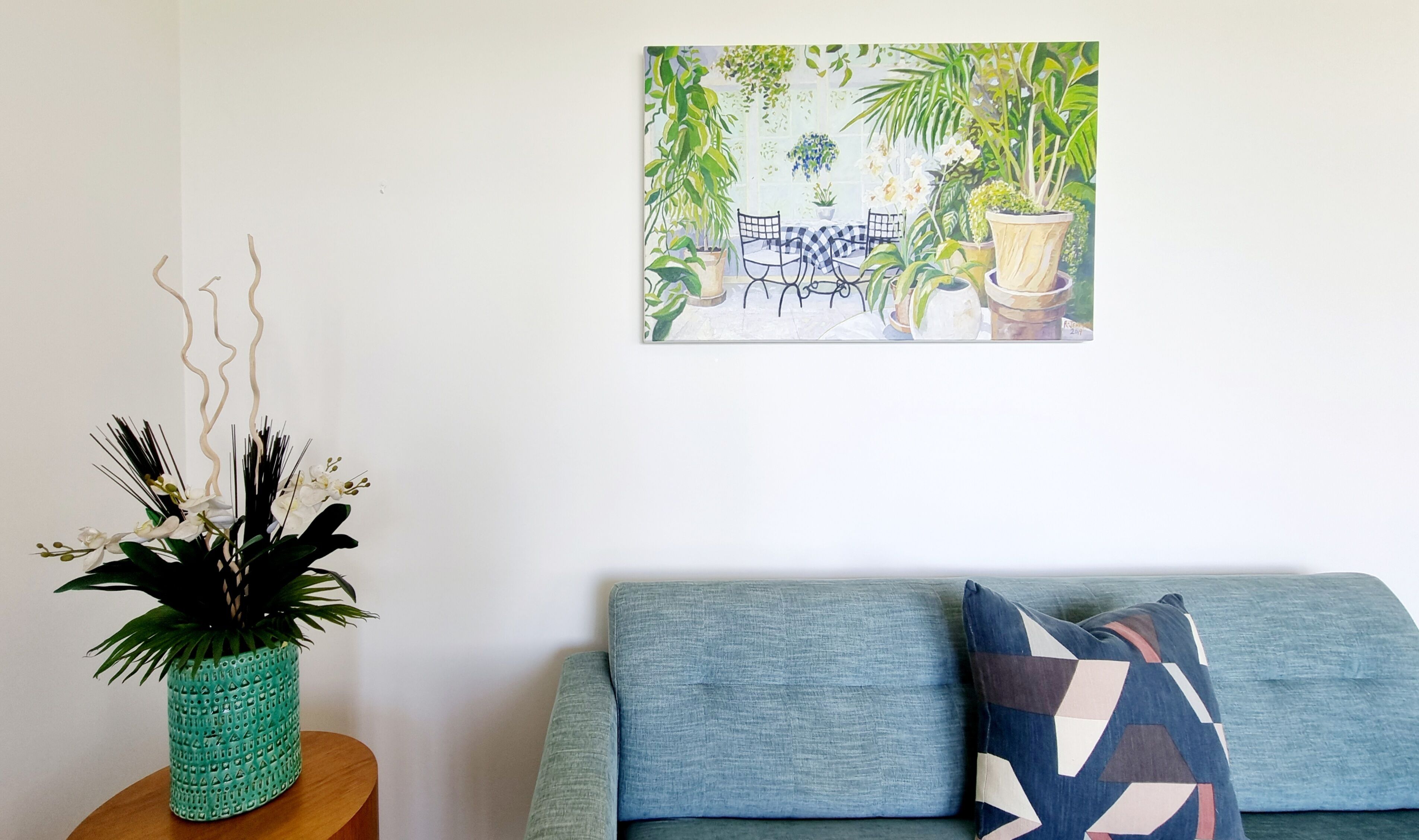 A living room with a blue sofa, a teal vase with white flowers, and a painting of a green outdoor scene.