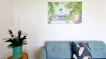 A living room with a blue sofa, a teal vase with white flowers, and a painting of a green outdoor scene.