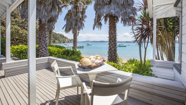 A covered outdoor seating area with a table set for two, overlooking a beach with palm trees and boats. - The Beach House (Kawau Island)