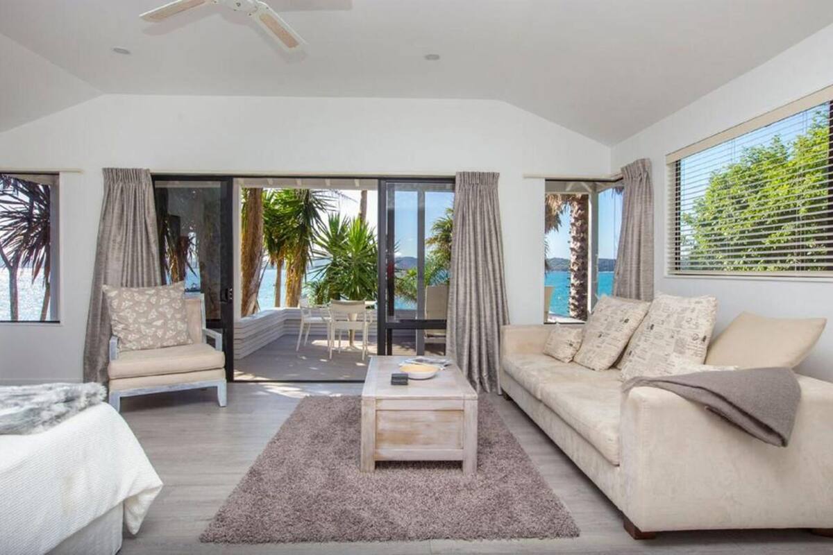A modern living room with a sofa, armchair, and coffee table, a balcony with a view of the sea, and a ceiling fan.