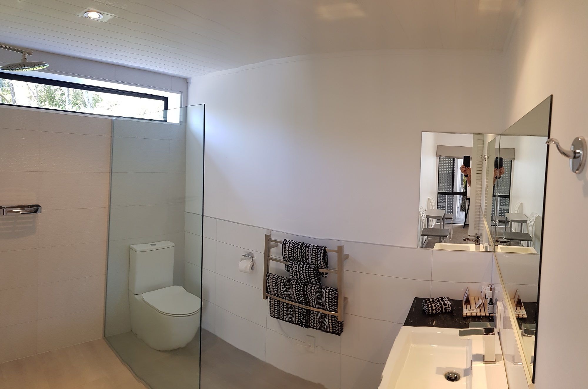 A modern bathroom with a glass shower enclosure, a toilet, a towel rack with patterned towels, a sink with a mirror, and a window providing natural light.