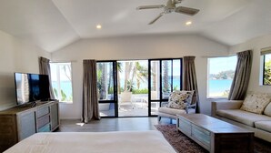A spacious bedroom with a large bed, a TV, a ceiling fan, and a view of the sea through large windows. - The Beach House (Kawau Island)