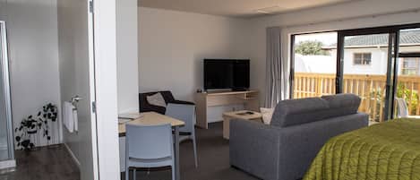 Ein Hotelzimmer mit Bett, grauem Sofa, Schreibtisch mit Stuhl, Fernseher und Balkon mit Holzlattenzaun.
