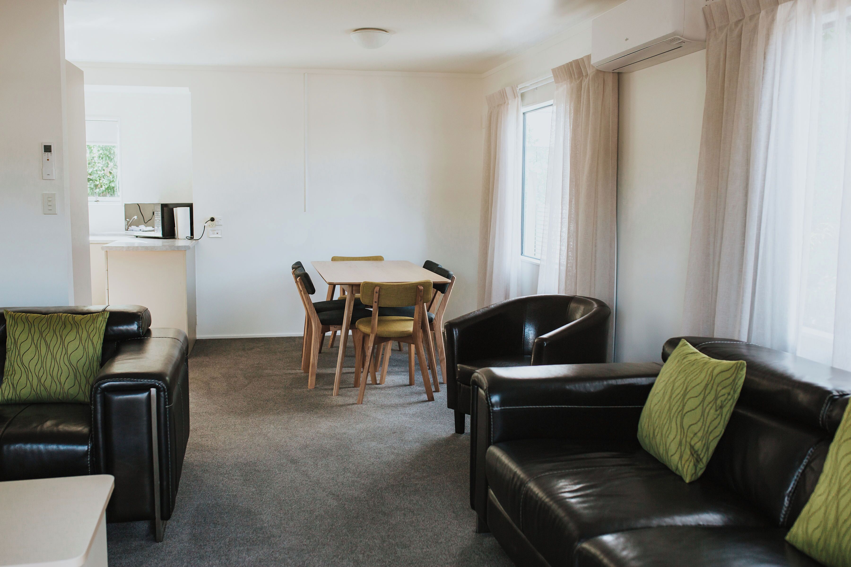 A living room with a black leather sofa, a dining table with chairs, and a window with curtains.