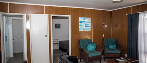 A room with a wooden paneled wall, two wicker armchairs with teal cushions, a black leather sofa, a wooden coffee table, and a bed in the background.
