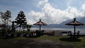 A coastal view with two umbrella tables and a seating area overlooking the sea, with mountains in the background. - Batur Lakeside Huts (Kintamani)