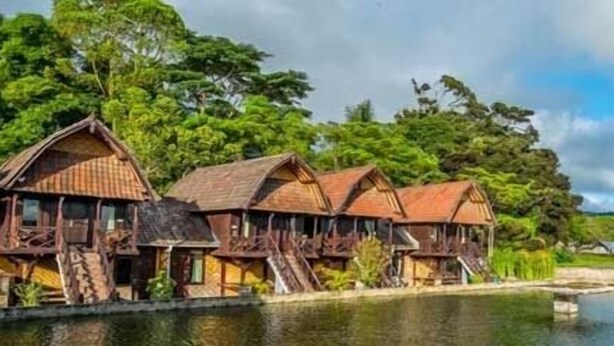 A row of wooden buildings with steeply pitched roofs, likely a lodge or resort, situated by a calm body of water with lush greenery in the background. - Batur Lakeside Huts (Kintamani)