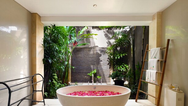 A large freestanding bathtub filled with rose petals, surrounded by lush greenery and a bamboo ladder with towels.