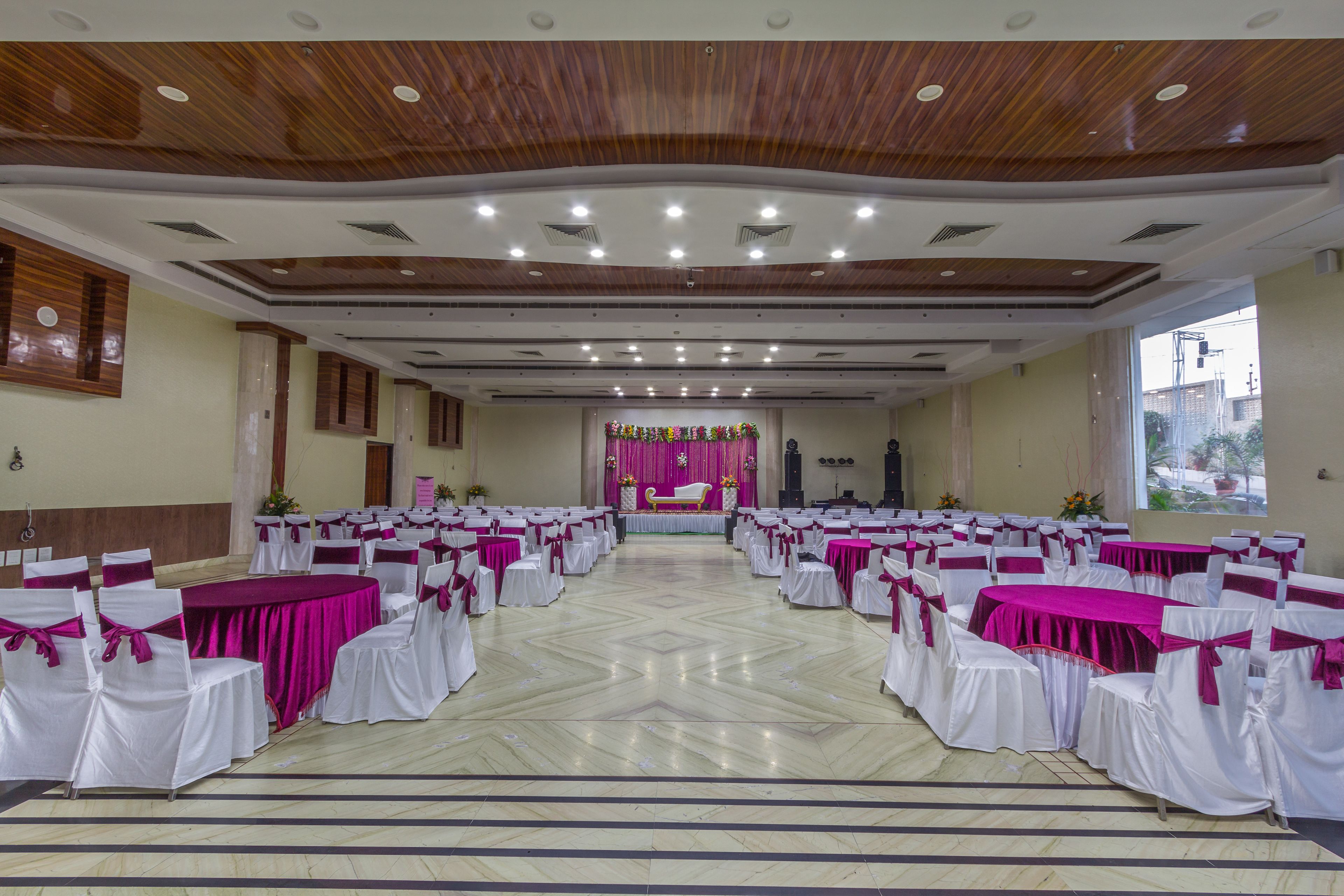 Ein Festsaal mit Reihen von Tischen und Stühlen, eine Bühne mit Blumenschmuck und eine Holzbalkendecke mit eingelassener Beleuchtung.