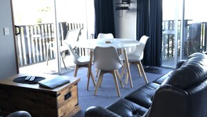 A modern living room with a leather sofa, a wooden coffee table, and a dining table with chairs. There is a sliding glass door leading to a balcony with a view of the outdoors. - The Kaka Point Motels (Kaka Point)