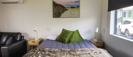 A bedroom with a bed, a chair, a nightstand, a lamp, a blanket, and a window with a view of a car outside.