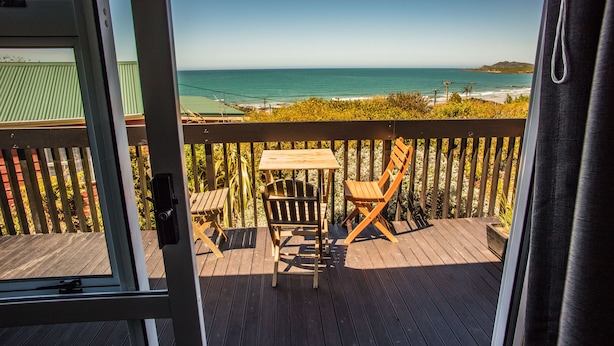 A wooden deck with a table and chairs, overlooking a beach with the ocean and a distant island. - The Kaka Point Motels (Kaka Point)