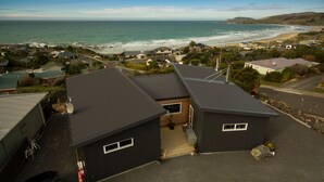 A residential area with houses near a beach, featuring a prominent view of the ocean and hills in the background. - The Kaka Point Motels (Kaka Point)