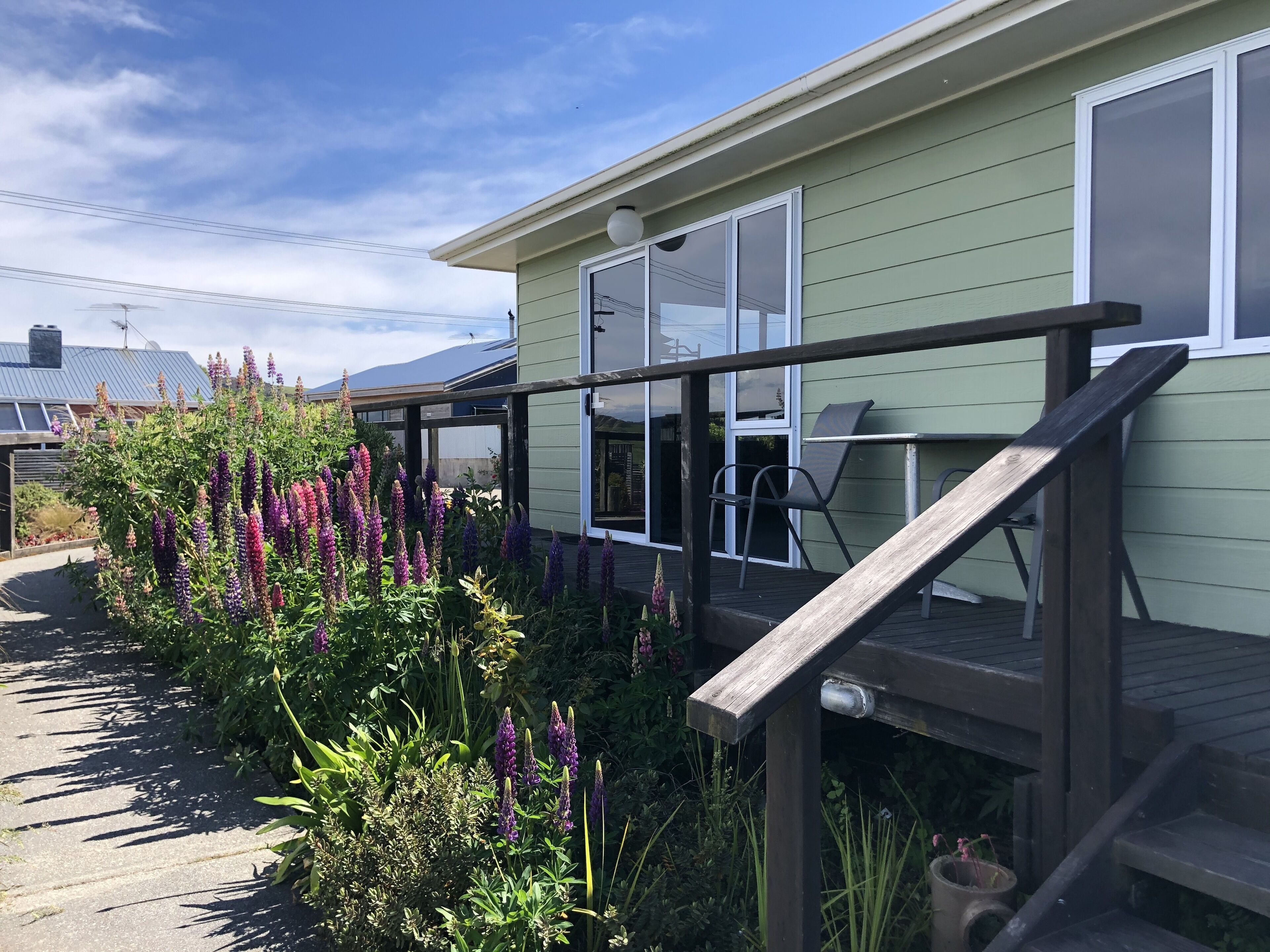 Una casa con terraza de madera, un espacio de estar cubierto al aire libre y un jardín con flores moradas.