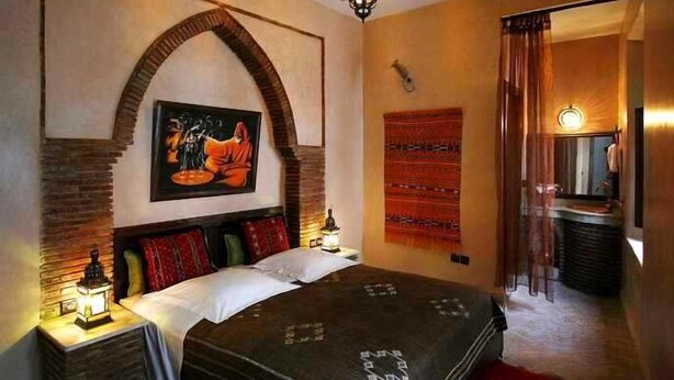Une chambre à coucher avec un grand lit, une table de chevet avec une lampe, un tableau accroché au mur et une tapisserie rouge à motifs.