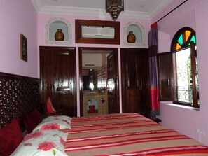 Un dormitorio con cama, cabecera de madera, un espejo, una ventana con cortinas y una puerta que da al baño.
