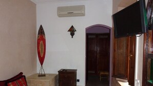 Une pièce comprenant un canapé, une petite armoire en bois, un climatiseur mural, une décoration murale et une porte en bois surmontée d’une arche violette.