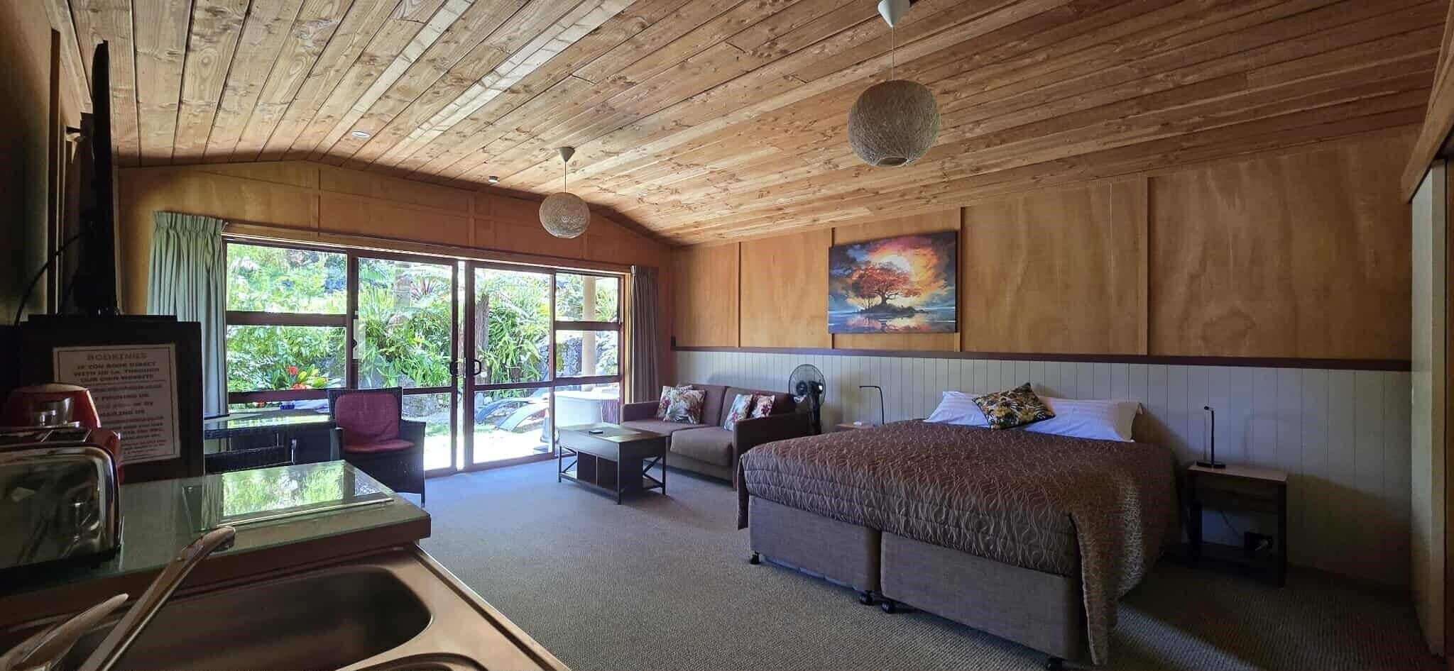 A hotel room with a large bed, a desk with a sink, a chair, a couch, and a large window offering a view of greenery.