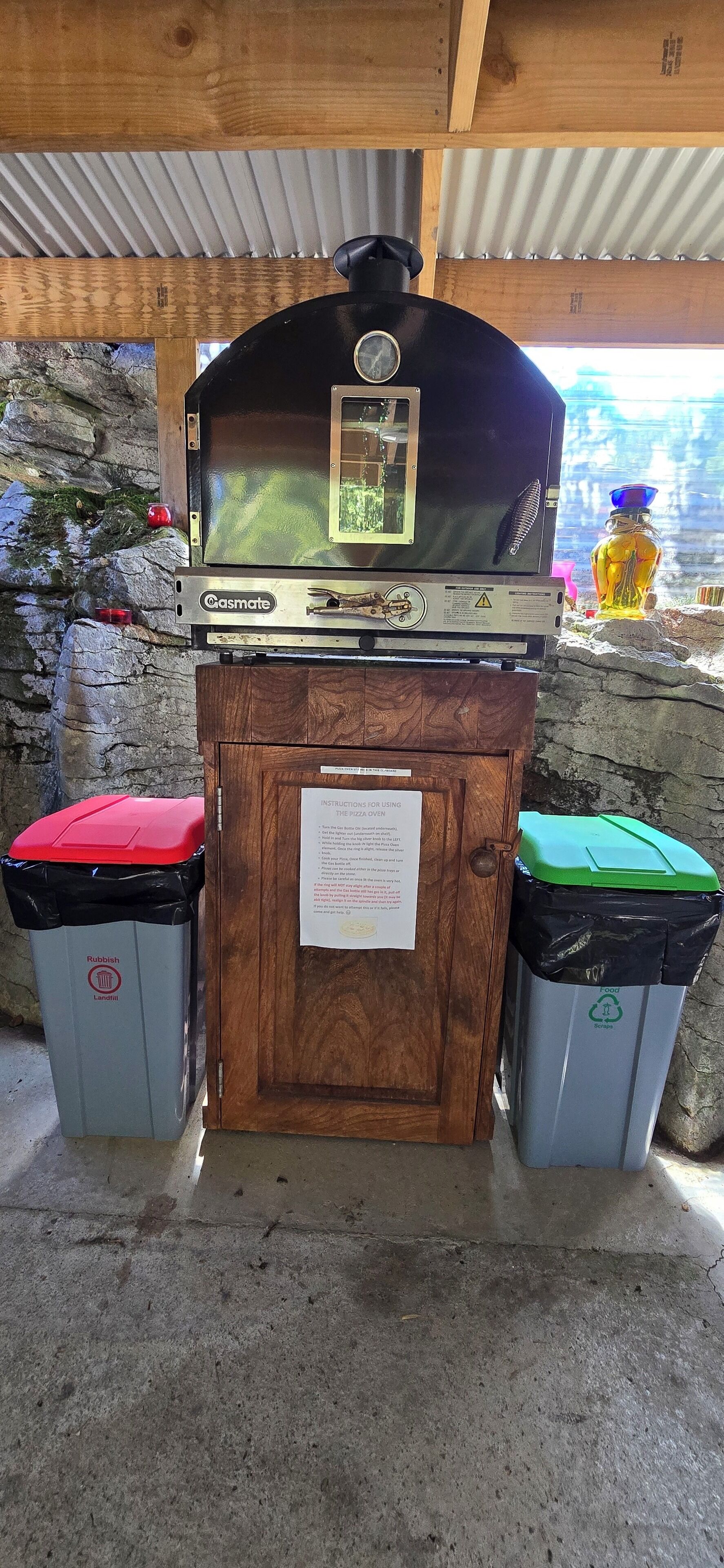 A pizza oven with a sign, two trash bins, and a stone wall in the background.