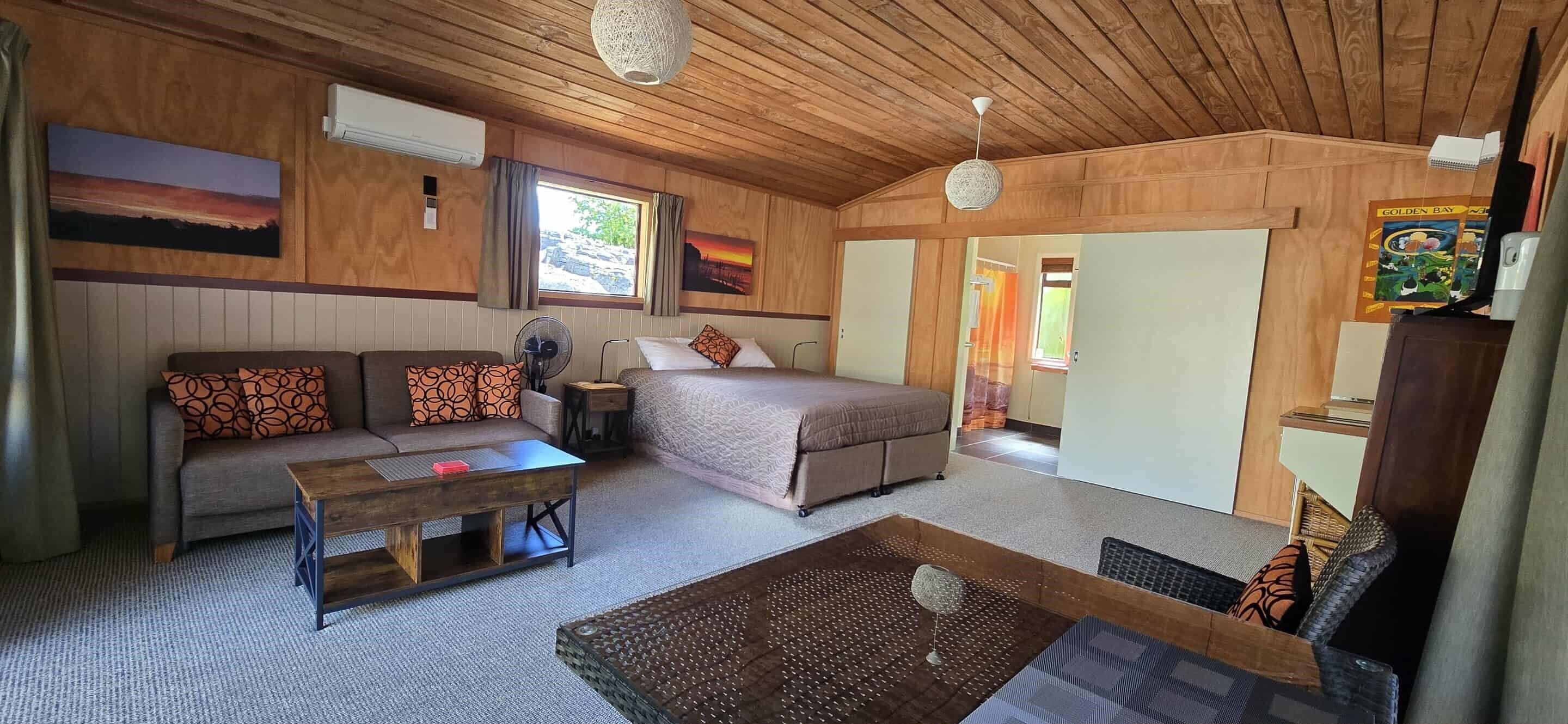 A cozy room with a bed, a sofa, a coffee table, and a fan. There are paintings on the walls and a window with curtains.