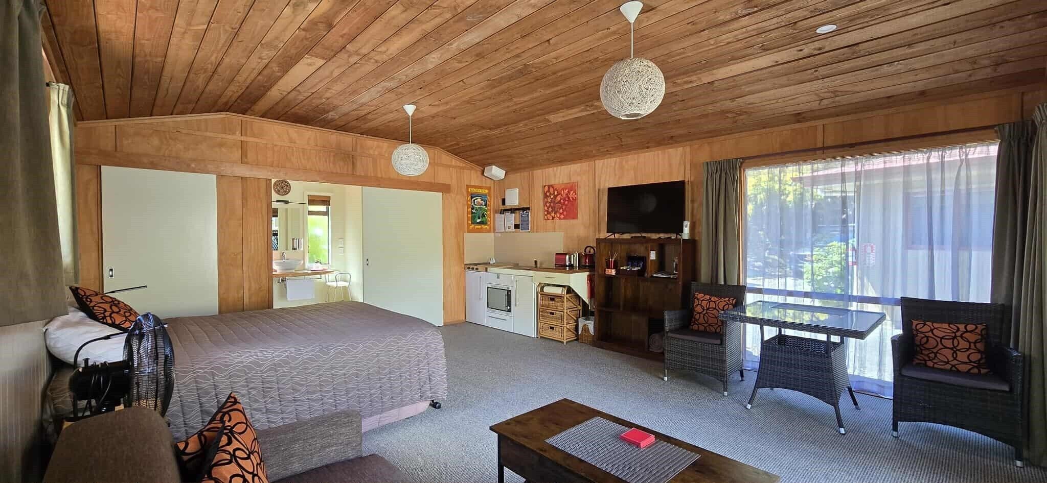 A cozy room with a bed, a small kitchenette, a dining area with wicker furniture, and a large window offering a view.