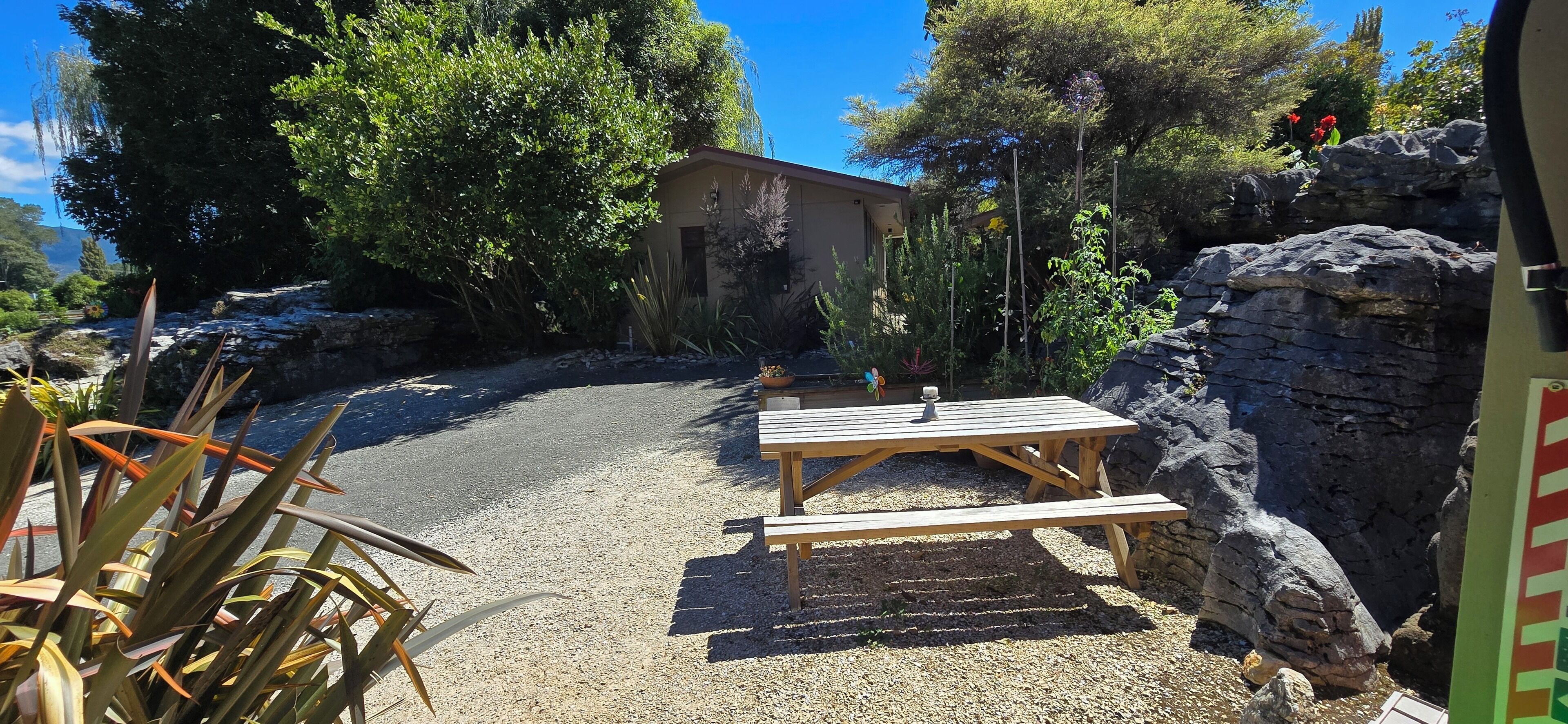 A gravel patio with a wooden picnic table and benches, surrounded by rocks and greenery.