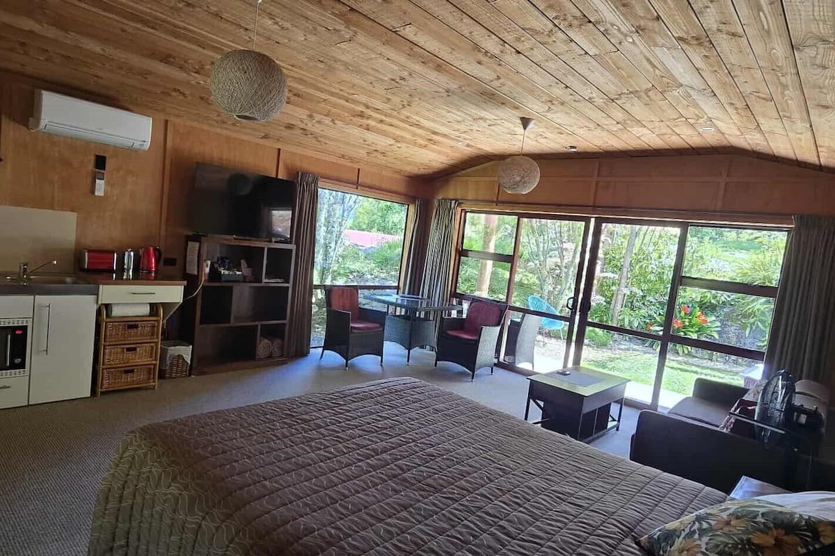 A cozy bedroom with a large bed, a kitchenette, a dining area with a table and chairs, and large windows offering a view of the outdoors.