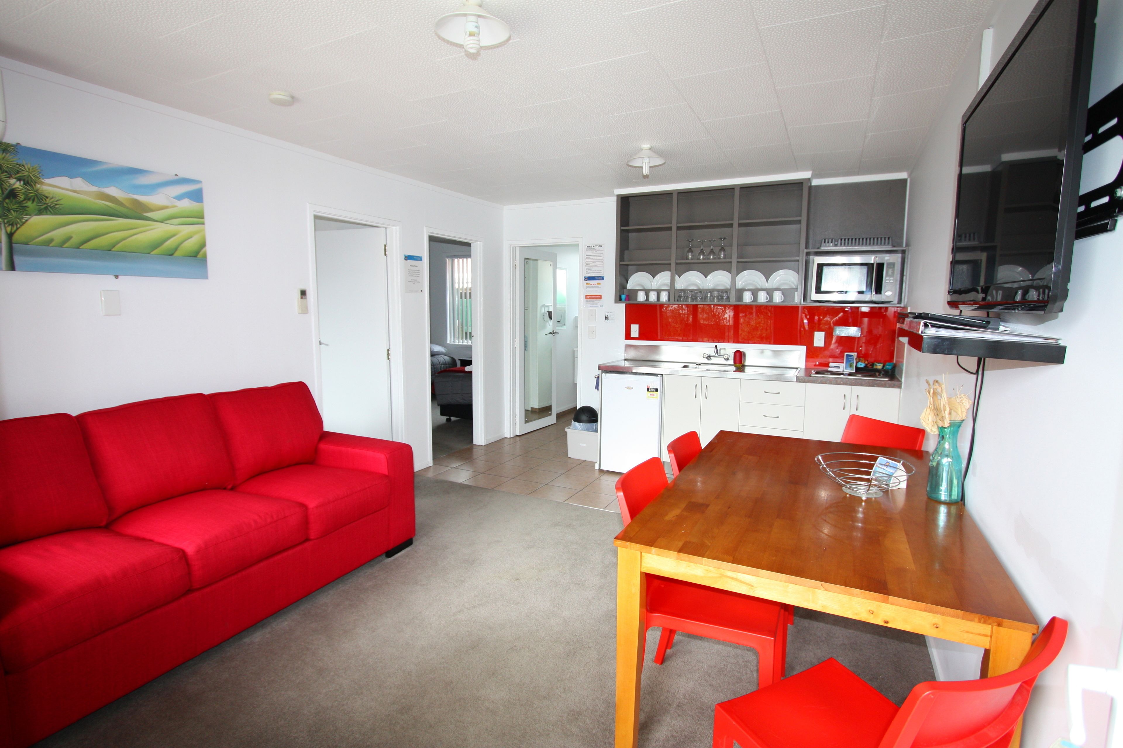 Una sala moderna con un sofá rojo, una mesa de comedor de madera con sillas rojas, una cocina con armarios blancos y un televisor de pantalla plana montado en la pared.