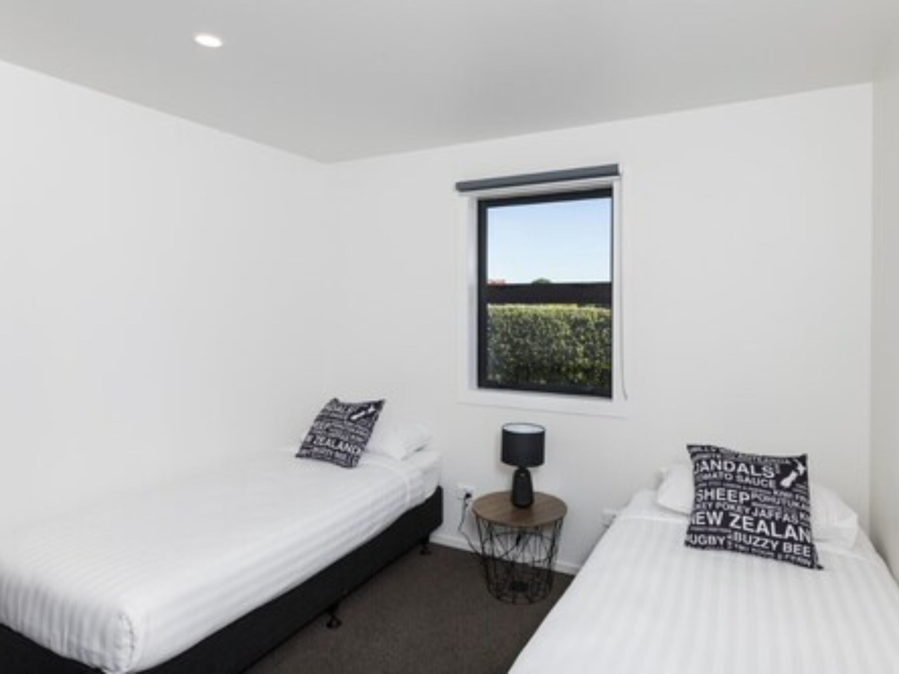 A bedroom with two single beds, a window with a view of greenery, a bedside table with a lamp, and decorative pillows.
