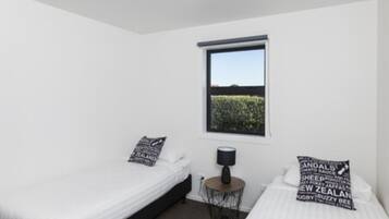 A bedroom with two single beds, a window with a view of greenery, a bedside table with a lamp, and decorative pillows.