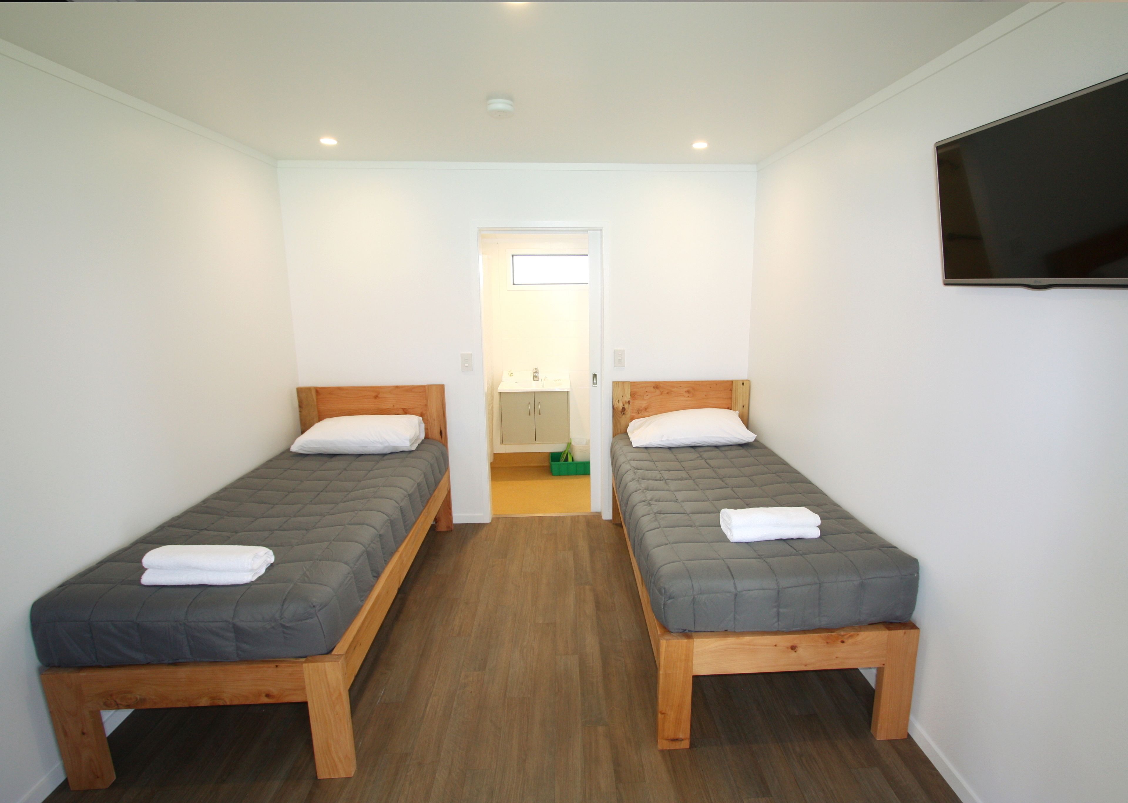 A room with two single beds, wooden headboards, a flat-screen TV mounted on the wall, and a small bathroom with a mirror and sink.