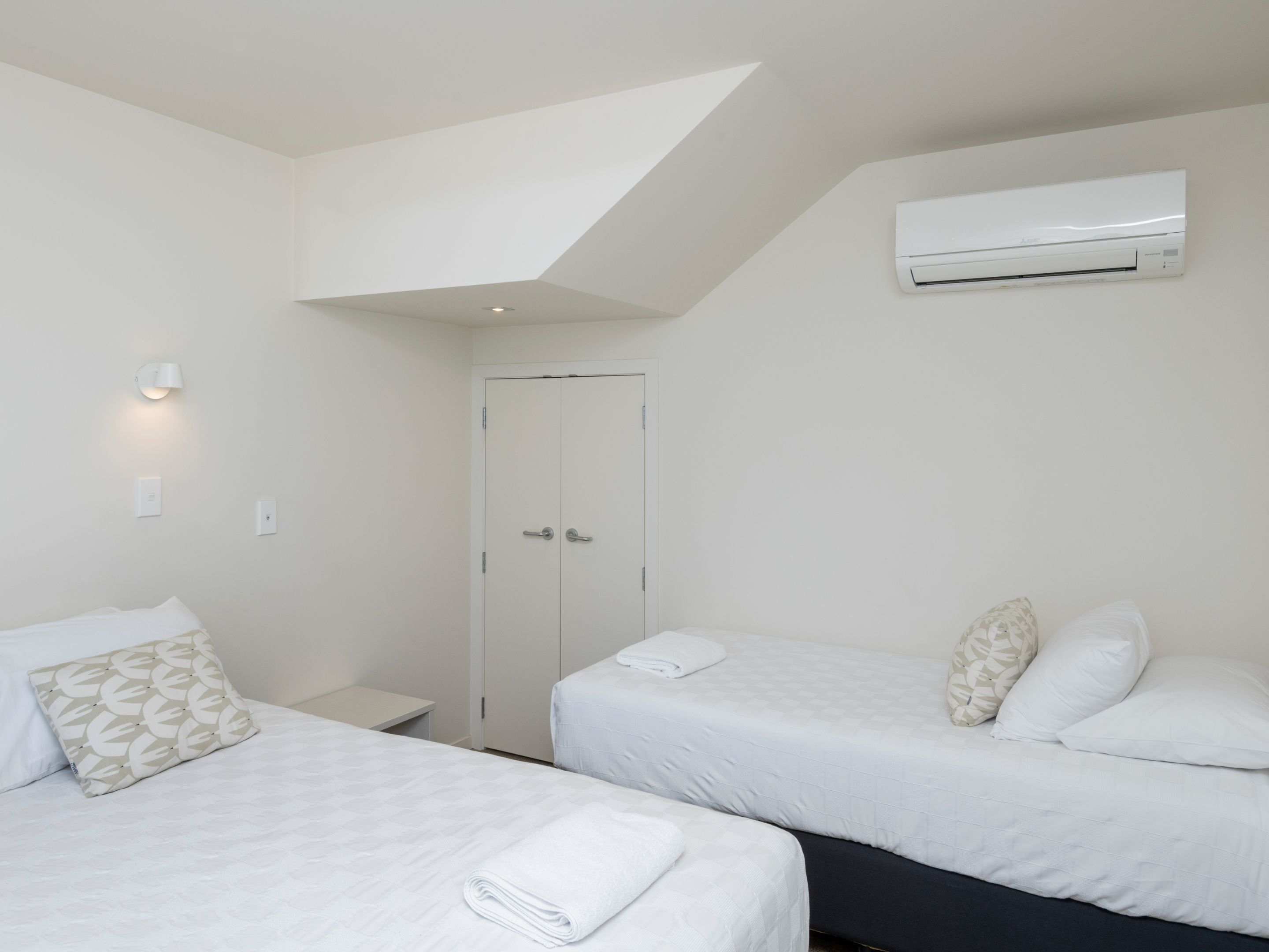 A hotel room with two beds, a wardrobe, an air conditioning unit, and a small bedside table.