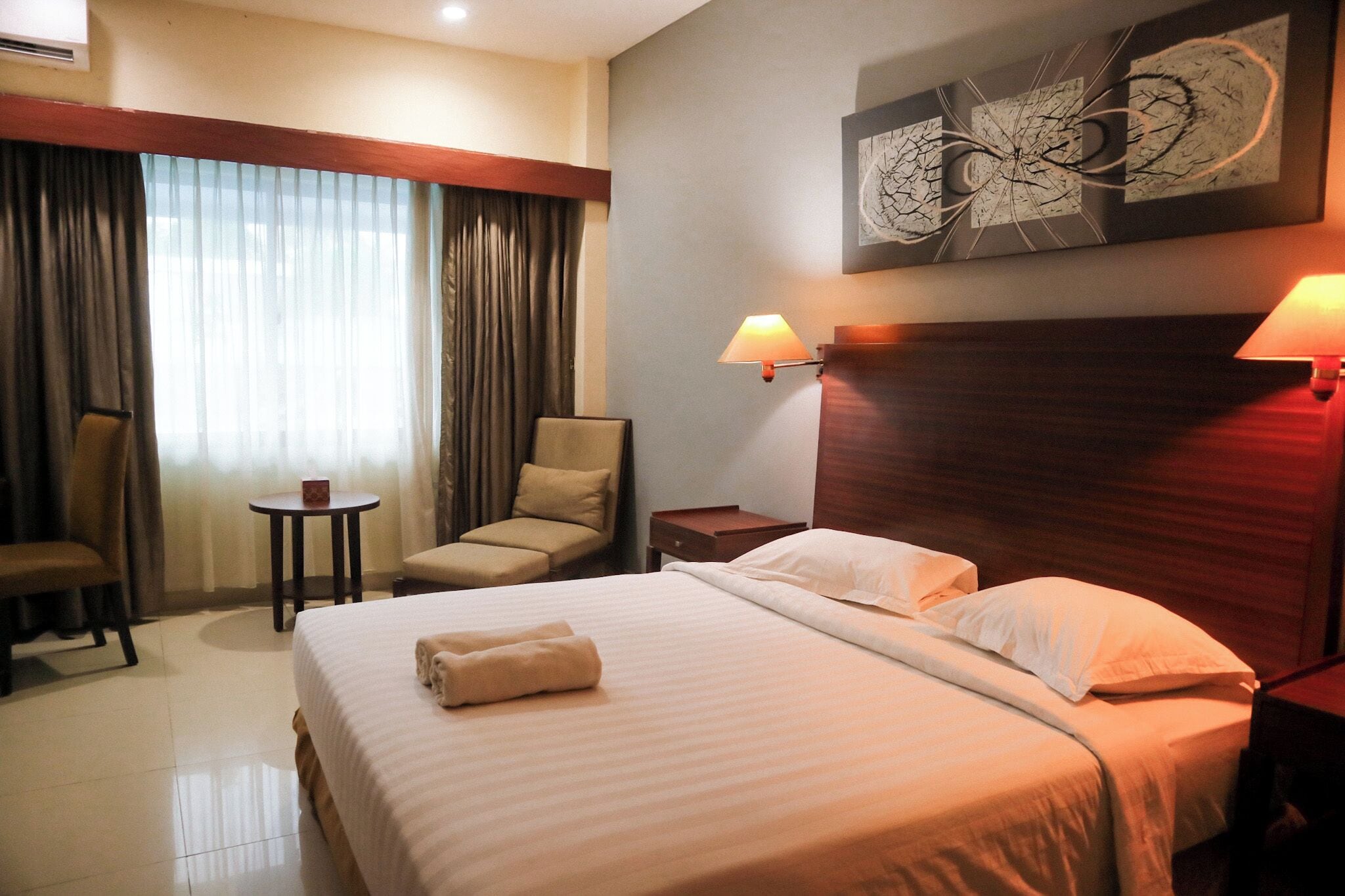A hotel room with a large bed, a chair, a small table, and a lamp.