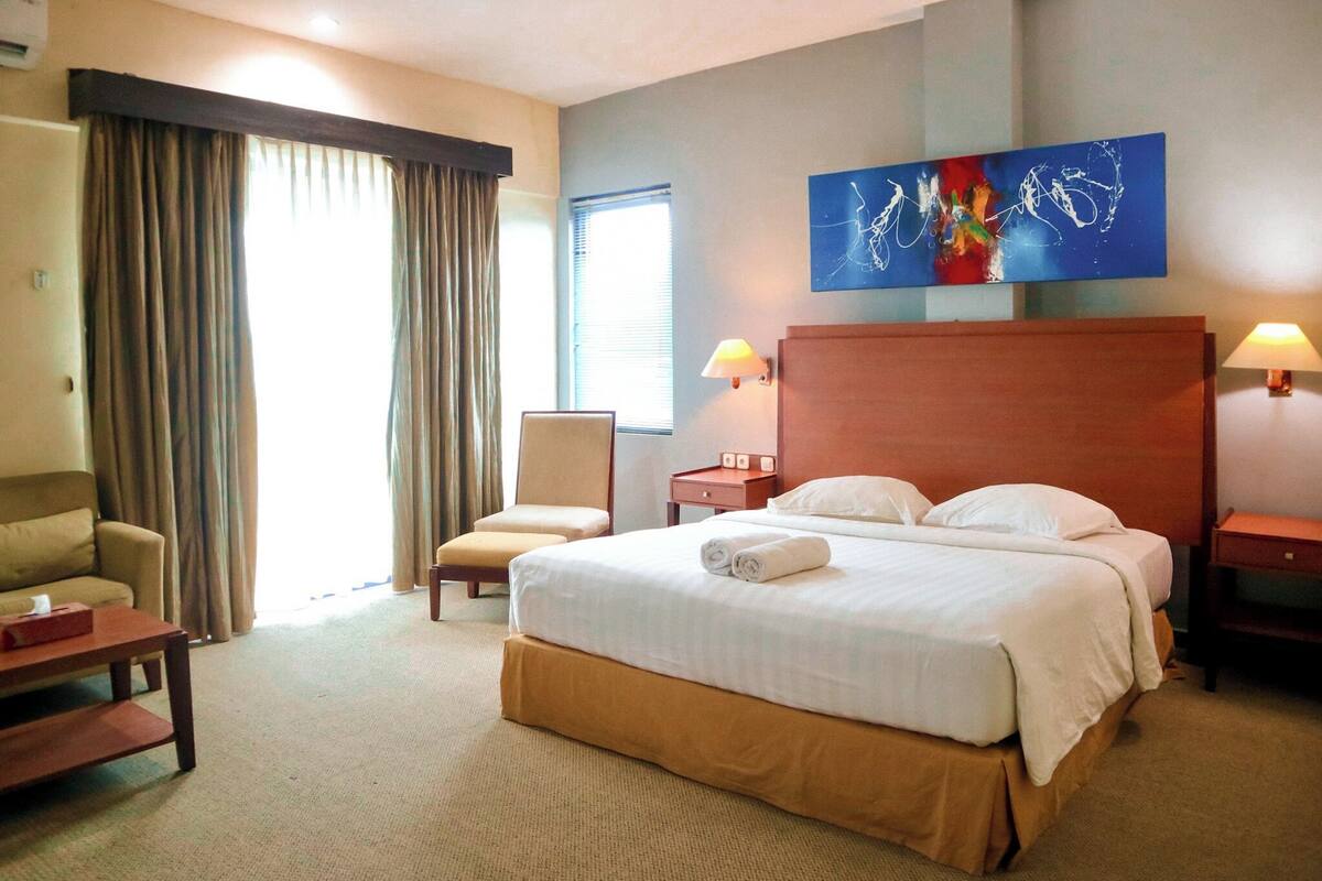 Hotellihuone, jossa on suuri sänky, tuoli, työpöytä ja ikkuna, jossa on verhot.