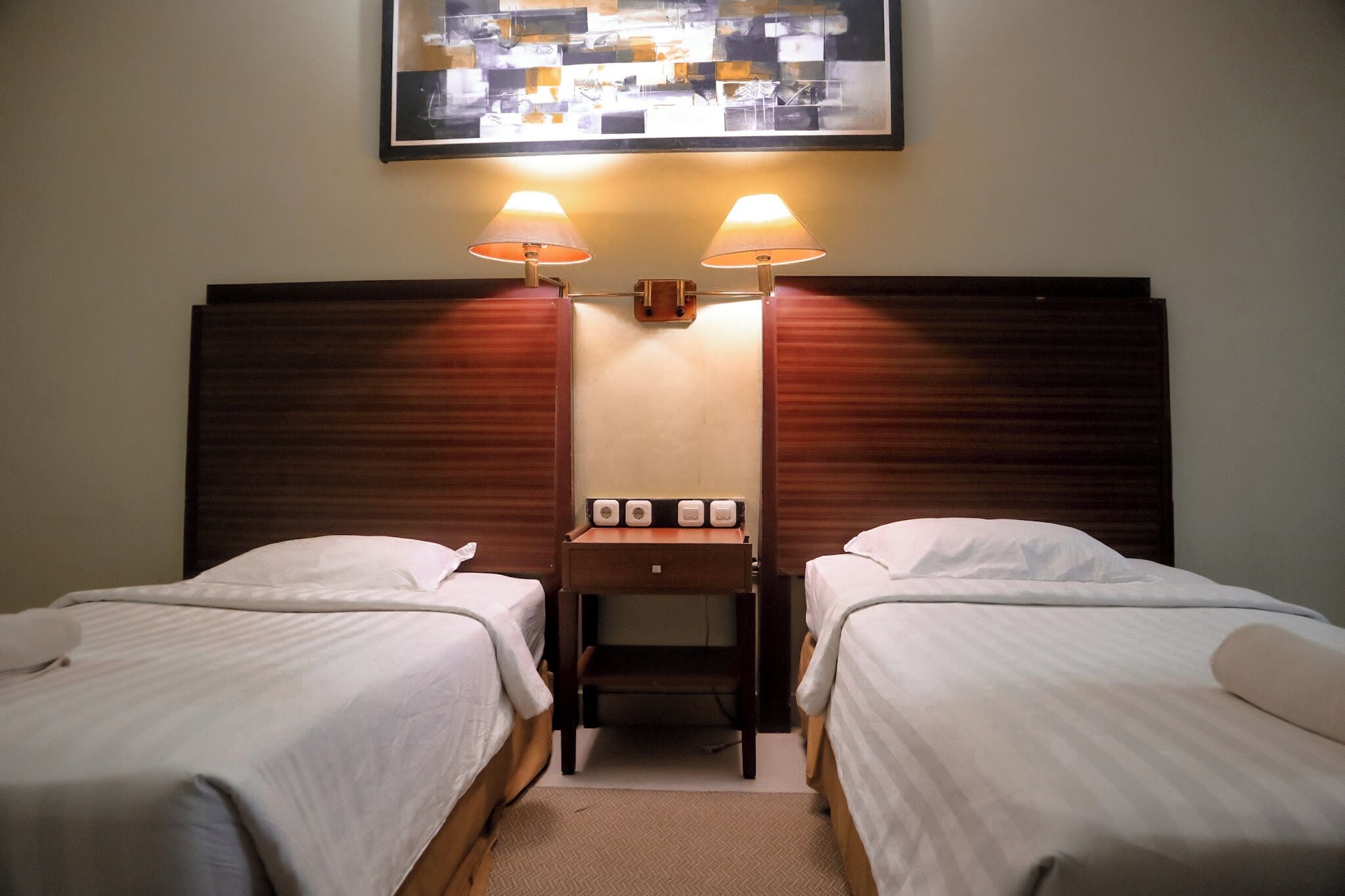 Habitación de hotel con dos camas individuales, una mesita de noche de madera con lámpara, y un cuadro enmarcado sobre la cama.