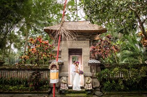 Una pareja está de pie frente a una construcción tradicional con techo de paja, rodeada de abundante vegetación y estatuas de piedra.