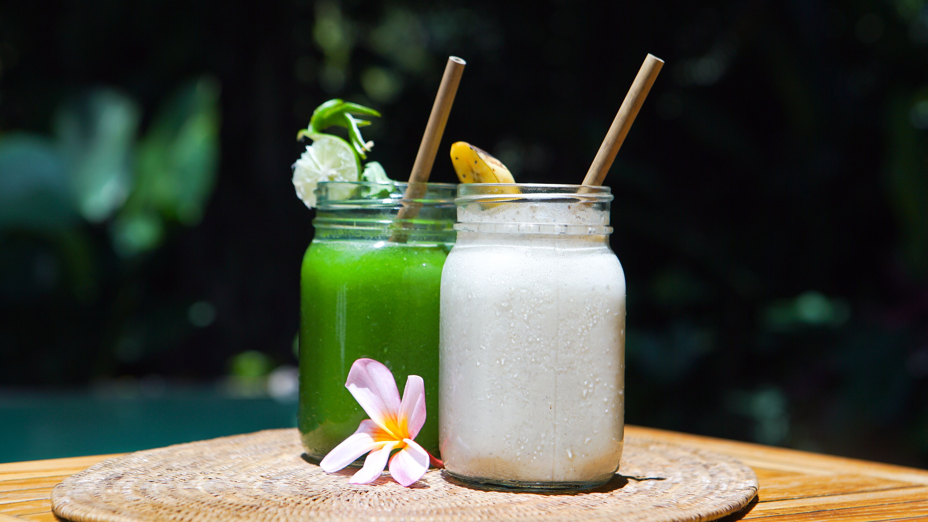 Due barattoli di vetro con cannucce su un tappetino intrecciato, uno verde e uno bianco, con un fiore accanto.