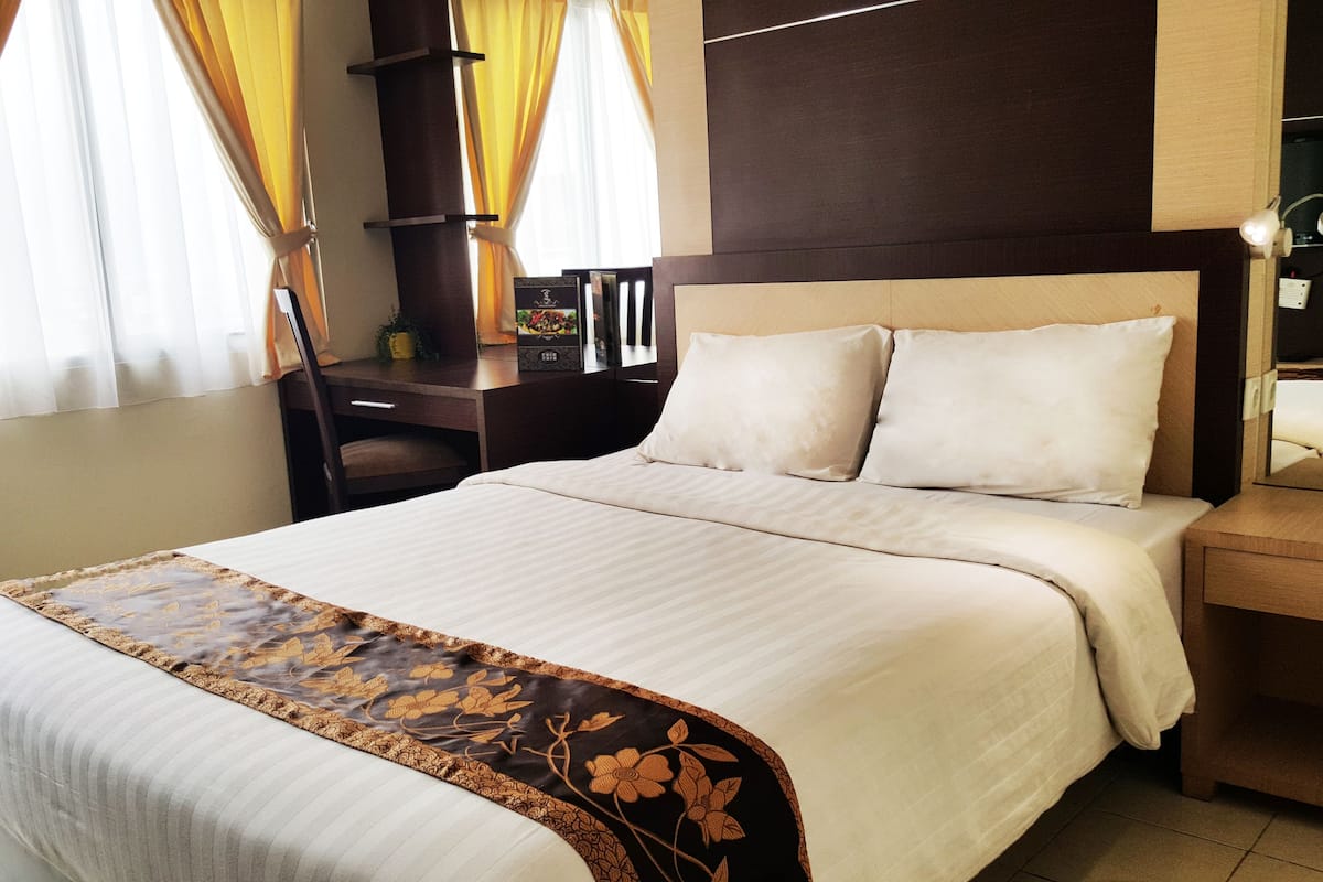 A neatly made bed with a floral bedspread, a wooden nightstand with a lamp, and a desk with a chair.