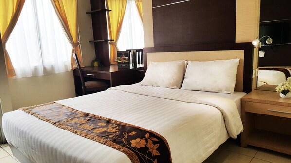A neatly made bed with a floral bedspread, a wooden nightstand with a lamp, and a desk with a chair. - Centro City Service Apartment (Jakarta)