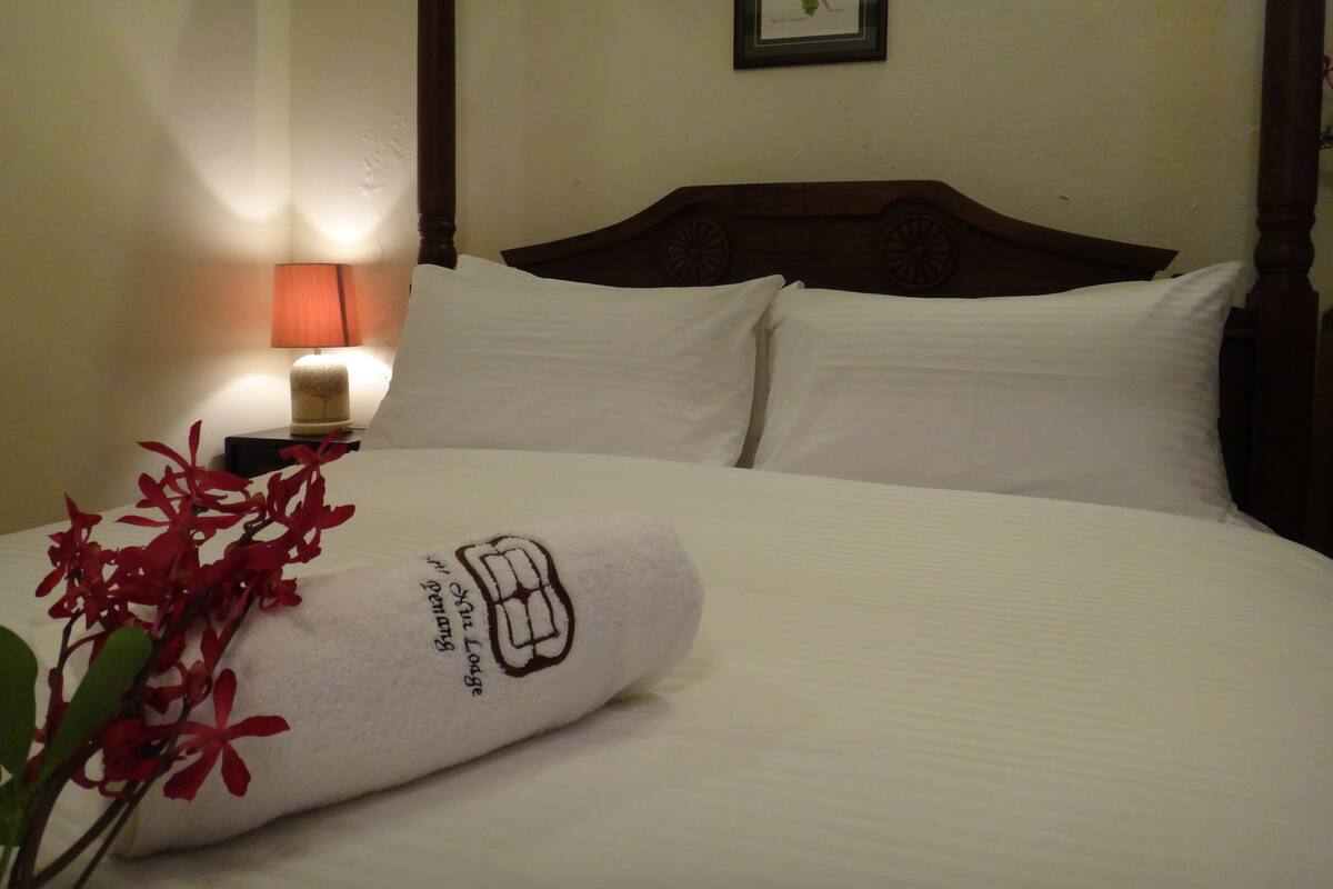 A neatly made bed with a white pillow and a towel with a logo, a wooden headboard, a wall-mounted picture frame, and a bedside lamp.